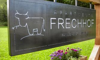 A sign with the inscription "Apartments Frechhof". Below it is a stylized cow depicted, and there are colorful flowers under the sign.
