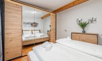 A modern bedroom with a cozy double bed and wooden paneling. The room is bright and tidy, with a large mirror and plants.