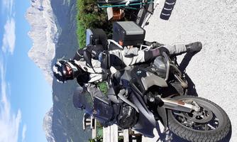 A motorcyclist in protective gear stands next to his motorcycle in a picturesque mountain landscape. In the background, tall mountains and a blue sky can be seen.