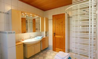 A modern bathroom with a large mirror, wooden bathroom furniture, and a shower. The walls are brightly tiled, and there are fresh towels on the floor.