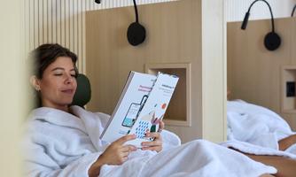 A woman in a bathrobe relaxes on a lounge chair and reads a book. The surroundings look modern and inviting.