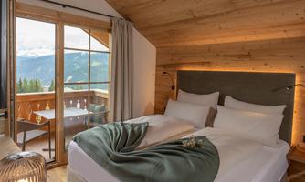 A cozy bedroom with wooden ceilings and warm lighting. The view overlooks a balcony with a mountain view.
