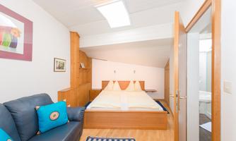 A cozy bedroom with a double bed and a blue sofa. The room has a bright wood design and a large window area.