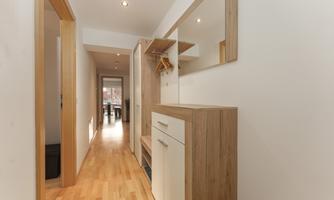 A bright hallway with hardwood floors and modern furniture. At the end of the hallway, access to another room is visible.