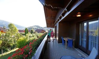 A spacious terrace with blooming flowers and a beautiful view of the mountains. Two people are enjoying the sun and the fresh air.