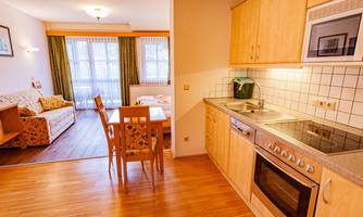 A bright and modern kitchen with wooden shelves and a table with chairs. In the background, there is a cozy living area with a sofa and large windows.