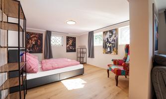 A modern bedroom with a double bed and a colorful armchair. Art pieces hang on the walls and large windows let in plenty of light.