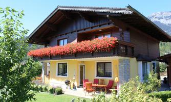A charming house with blooming balcony flowers and a beautiful garden. In the background, tall mountains can be seen.