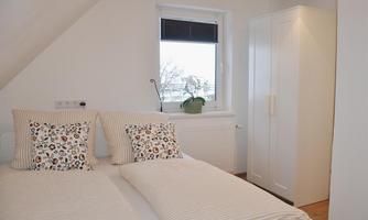 A cozy bedroom with two single beds and floral patterned cushions. Against the wall stands a white wardrobe and a window with a view outside.