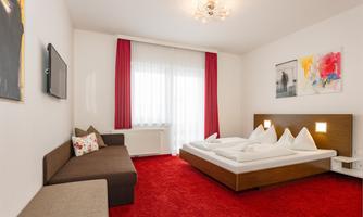 A modern room with a red carpet, a cozy sofa, and a large double bed. Bright curtains provide plenty of light.