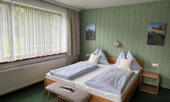 A cozy bedroom with a double bed, two pillows, and a small table. Large windows provide ample daylight and a relaxed atmosphere.