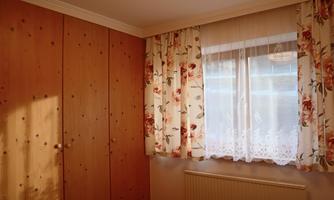 A cozy bedroom with wooden wardrobes and a light curtain in floral patterns. The window lets in natural light and creates an inviting atmosphere.