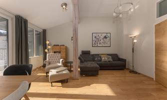 A modern living room with wooden floors and large windows. It is brightly furnished with a cozy sofa, an armchair, and a piece of artwork on the wall.