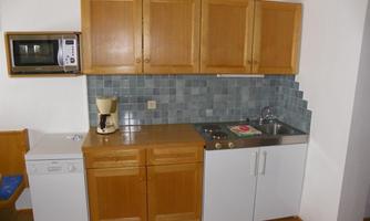 A small kitchen with wooden cabinets and light walls. It is equipped with a stove, a sink, a coffee maker, and a refrigerator.