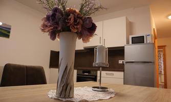 A cozy dining area with a table, a vase with colorful flowers, and a candle. In the background, a modern kitchen and a refrigerator are visible.