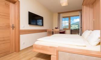 A modern bedroom with a double bed and wooden-colored furniture. The room has a window with a view of the mountains and a television on the wall.