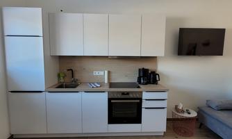 A modern kitchen with white cabinets and a cooking area. Next to the kitchen is a gray couch and a television on the wall.
