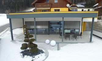 A modern building with large windows in a snowy landscape. In front of the house, there is a table with chairs and decorative plants.