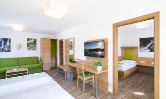 A modern room with a double bed and a single bed. The decor is bright and friendly, featuring a green sofa area and a television.