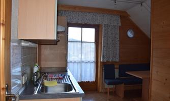 A small kitchen with wooden furniture and a dining area. Next to a window, there is a clock on the wall.