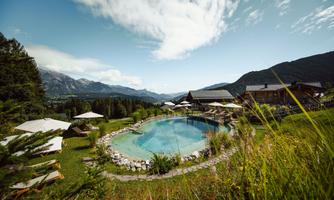 A beautiful pool surrounded by green grass and mountains in the background. The relaxed atmosphere is enhanced by sun umbrellas and loungers.