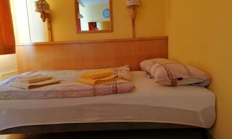 A simple bedroom with a bed, pillows, and additional towels. The walls are painted yellow and there is a mirror in the room.