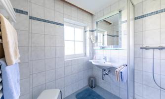 A modern bathroom with white tiles and a shower. There is a sink, a mirror, and towels in soft colors.