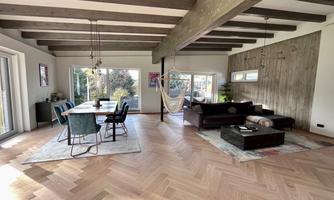 A modern living room with a wooden beam ceiling and large windows. The room is furnished with a sofa, a table, and a hammock.