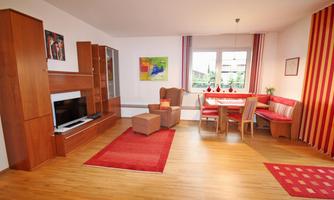 A bright living room with wooden floors and large windows. It is furnished with a cozy armchair, a dining table, and a TV unit.