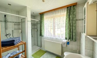 A bright bathroom with a shower and a large window front. Towels are neatly arranged on a wooden shelf, and green curtains provide a great view outside.