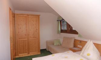 A small, cozy bedroom with a single bed and a sofa. The room has wooden furniture and windows that let in natural light.