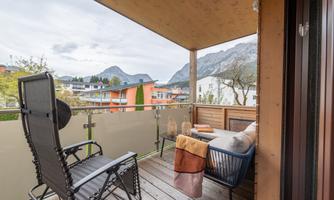 A cozy balcony with a comfortable chair and a sofa. In the background, mountains and a quiet residential area can be seen.