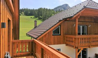 A wooden house with a balcony overlooks a green meadow and wooded hills. The architecture is rustic and inviting.