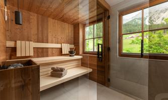 A modern wooden sauna with a relaxation bench and a small bucket. The view from the window shows a green mountain landscape.
