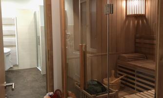 A modern sauna area with wood paneling and a glass door. In the background, a bathroom with white tiles is visible.