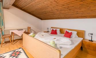 A cozy bedroom with a wooden ceiling and a large bed. The decoration includes red cushions and a small table with chairs.