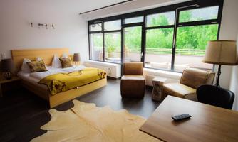 A modern bedroom with a large bed and wooden furniture. Large windows offer a view of greenery.