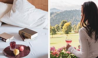A tray with a glass of juice, an apple, and a pear lies on a bed next to two books. A woman is looking at a picturesque landscape with trees and blooming flowers while holding a glass with a pink drink in her hand.