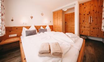 A cozy bedroom with a large bed and wood paneling. The windows are decorated with floral curtains, and there are soft towels on the bed.