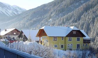 A picturesque mountain village in winter with snow-covered houses. The surroundings are characterized by tall, forested mountains and a clear sky.
