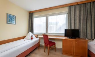 A simple room with two beds and a table. There are large windows with curtains and a television on a piece of furniture.