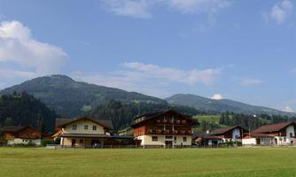 A picturesque village with traditional houses and gentle hills in the background. The sky is blue and there is a green meadow in the foreground.