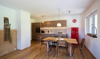 A modern kitchen with a large wooden dining table and stylish chairs. In the background, there is a red refrigerator, and there are natural light sources.