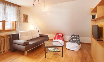 A cozy living room with a gray sofa, two bean bags in red and blue, and a glass table. The rooms are bright and friendly with wooden flooring and large windows.