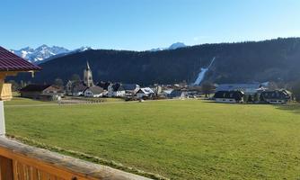 A picturesque landscape featuring a small village, surrounded by mountains and a green meadow. In the background, a waterfall is visible, and the sky is clear and blue.