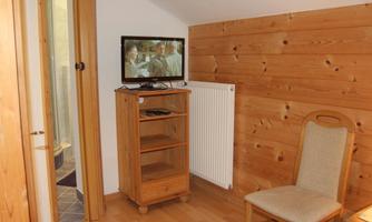 A simple room with a wooden shelf, a television on the wall, and a chair. The walls are made of wood and there is a radiator.