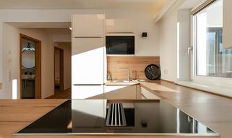 A modern kitchen with bright wooden surfaces and a cooktop in the center. The room has large windows that let in plenty of natural light.