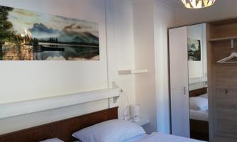 A cozy bedroom with a large mirror and a double bed. A landscape painting with mountains and a lake hangs on the wall.