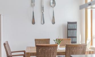 A modern dining room with a wooden table and woven chairs. Large decorative utensils hang on the wall.