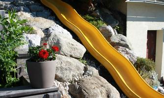 A yellow slide that stands on a slope next to a building. Around the slide are stones and a flower pot with red flowers.